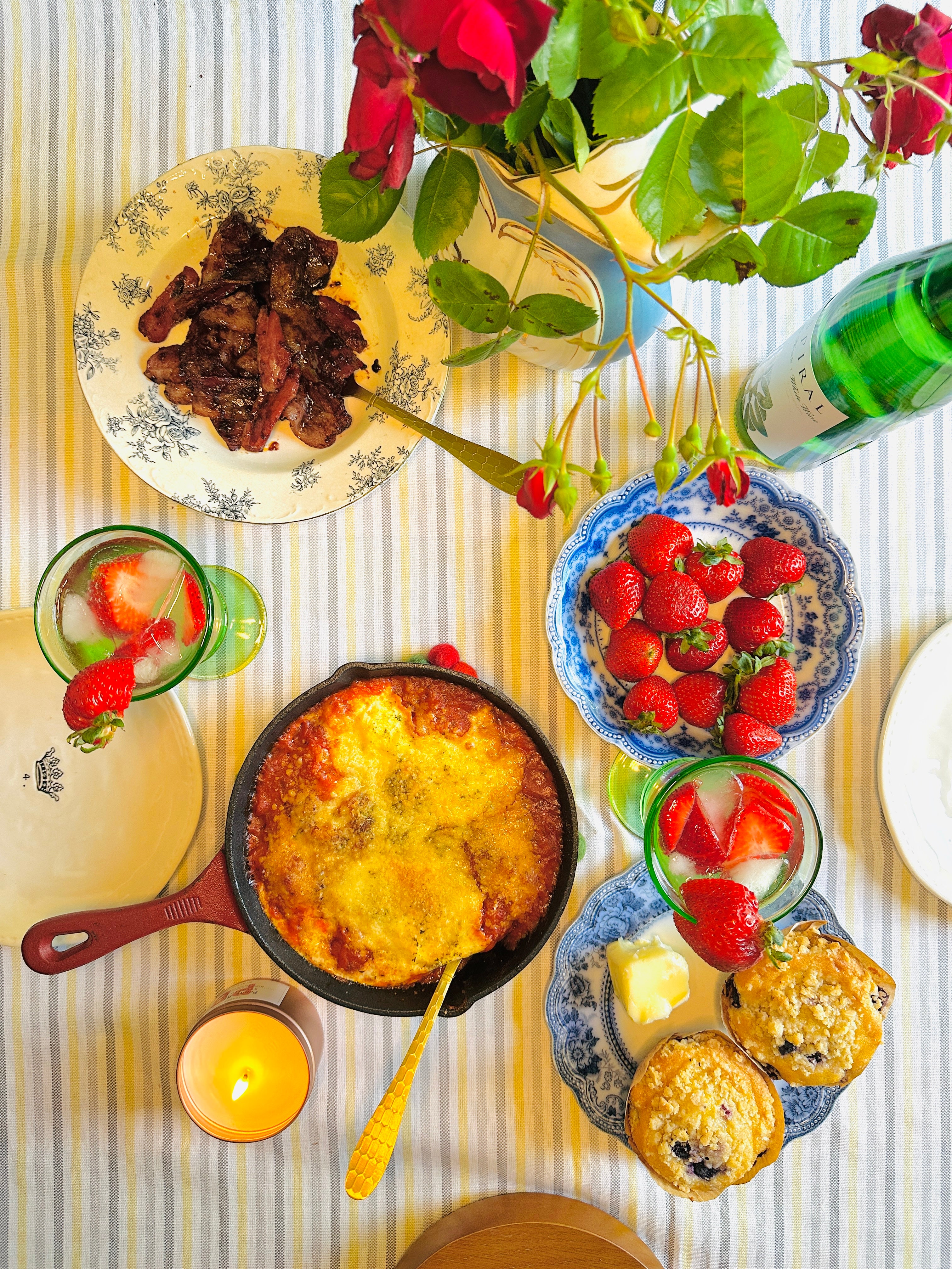 Dining table with a variety of food including a skillet, plates with strawberries, and a bottle of wine.