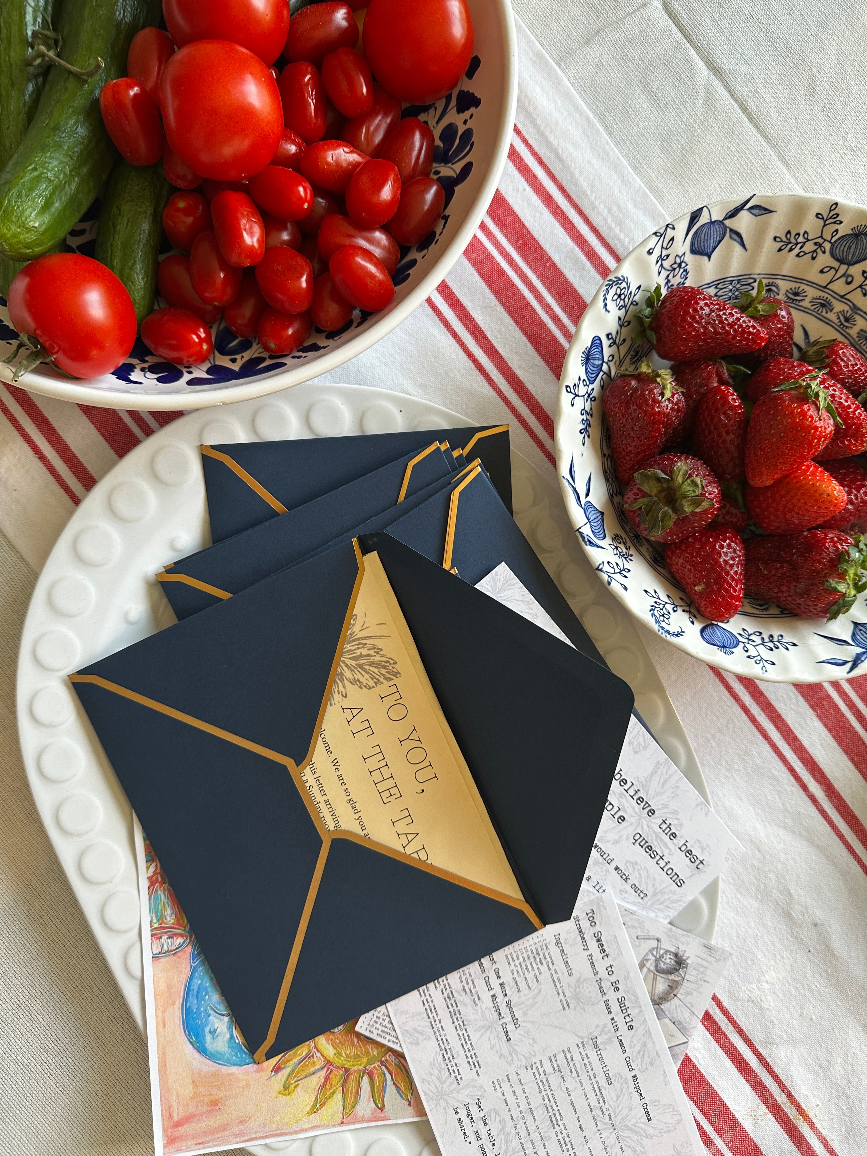Navy blue and gold envelopes on a table with bowls of tomatoes and strawberries.