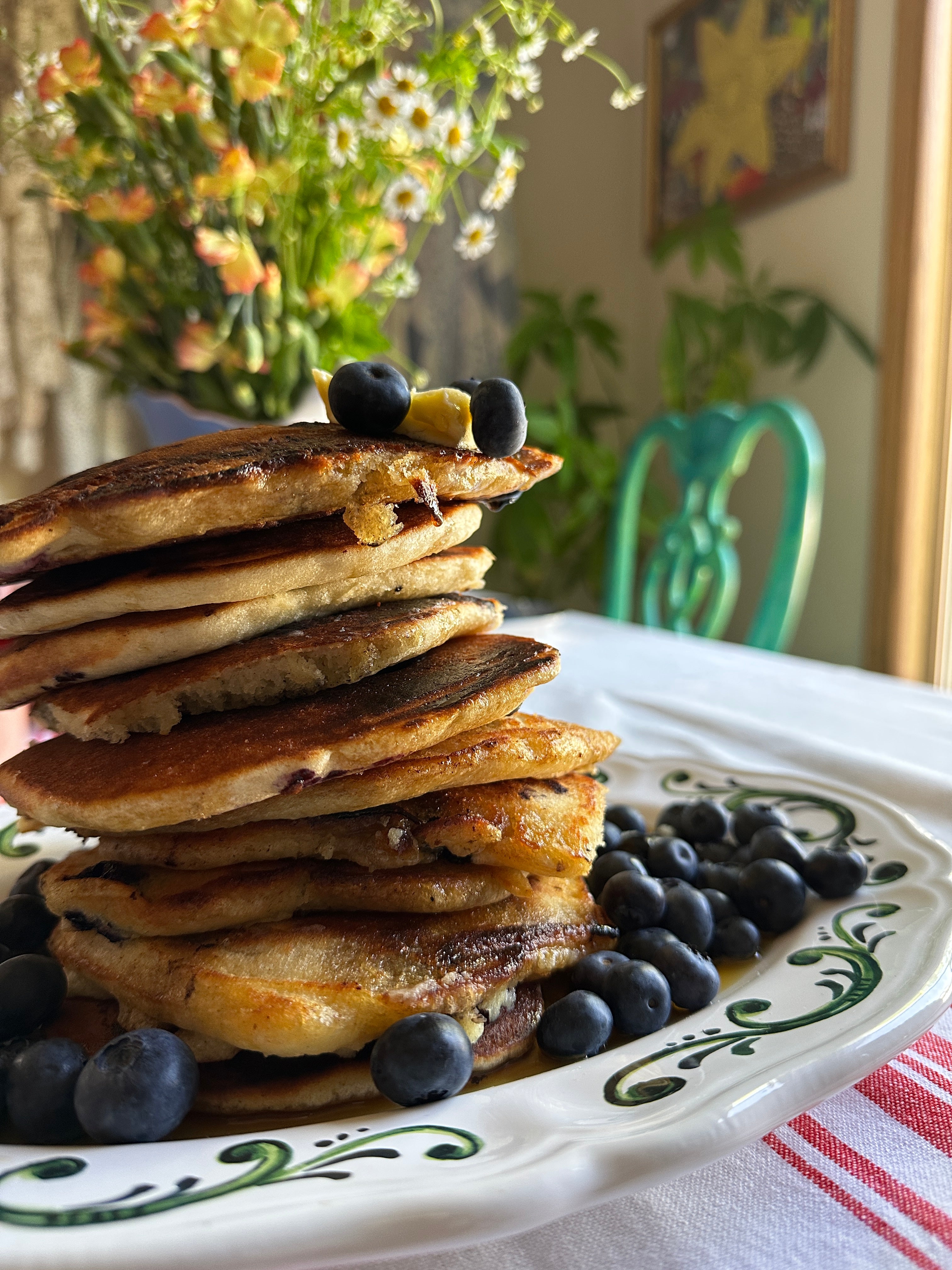 Stack of pancakes with blueberries on a decorative plate, with a blurred background of flowers and furniture.