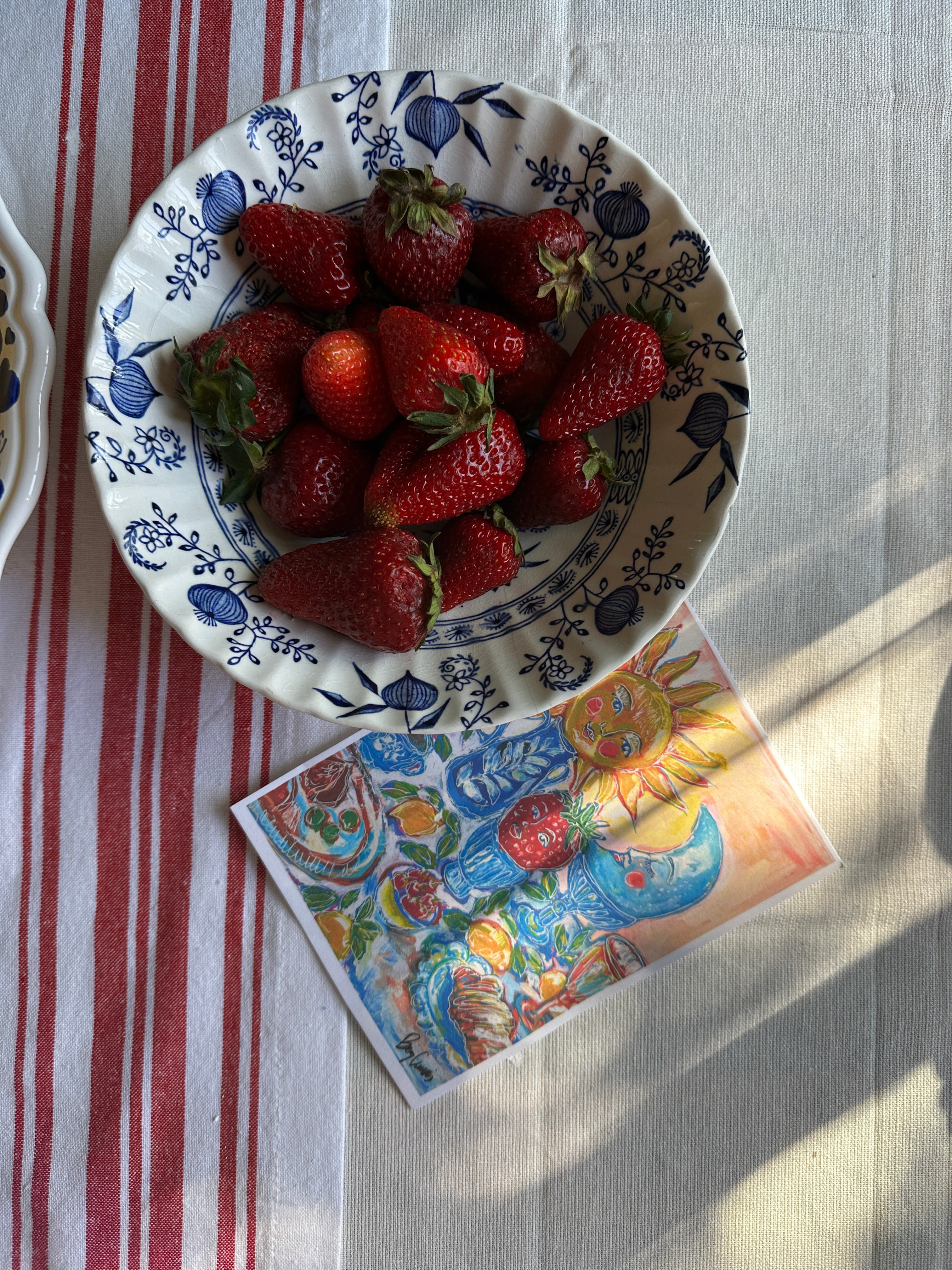 Bowl of strawberries on a patterned plate with a colorful card beside it on a striped tablecloth.